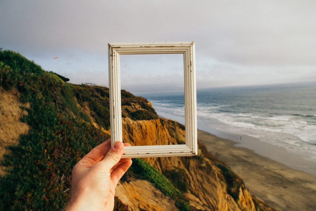 the bigger picture, a picture showing a human hand holding a frame through and outside which a beach can be seen